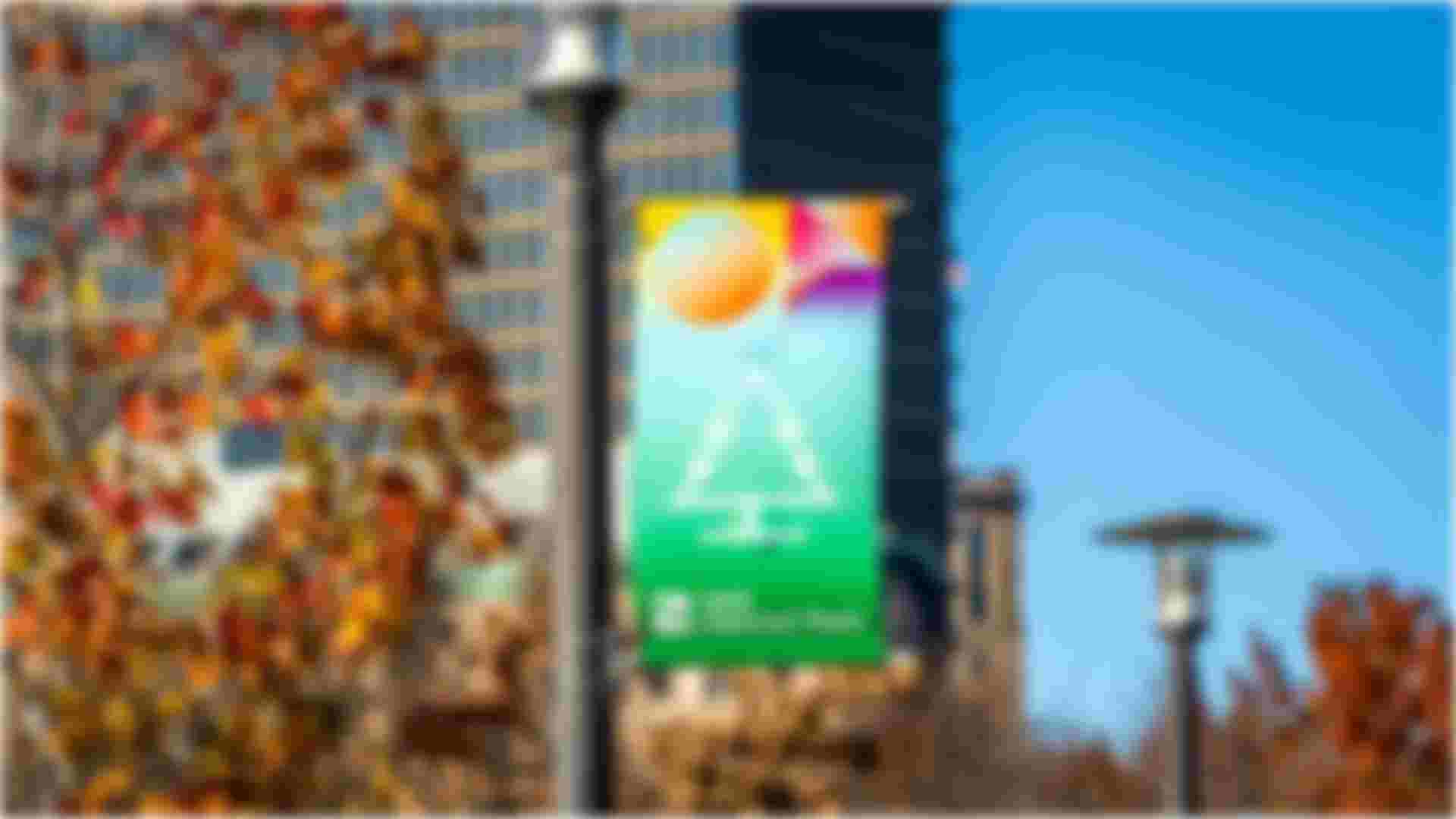 A colorful banner featuring a Christmas tree icon hanging from a lamppost at 195 District Park. The background shows autumn leaves with vibrant orange and red hues, and tall buildings in the distance against a clear blue sky.