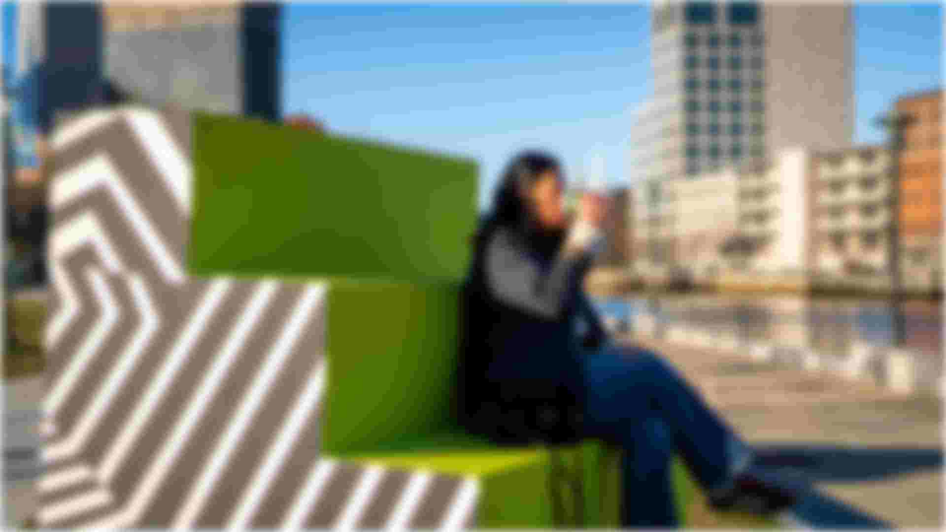 A woman sitting on a green STEWP! drinking a coffee with a geometric black and white pattern on the side of the steps and the Providence River in the background.