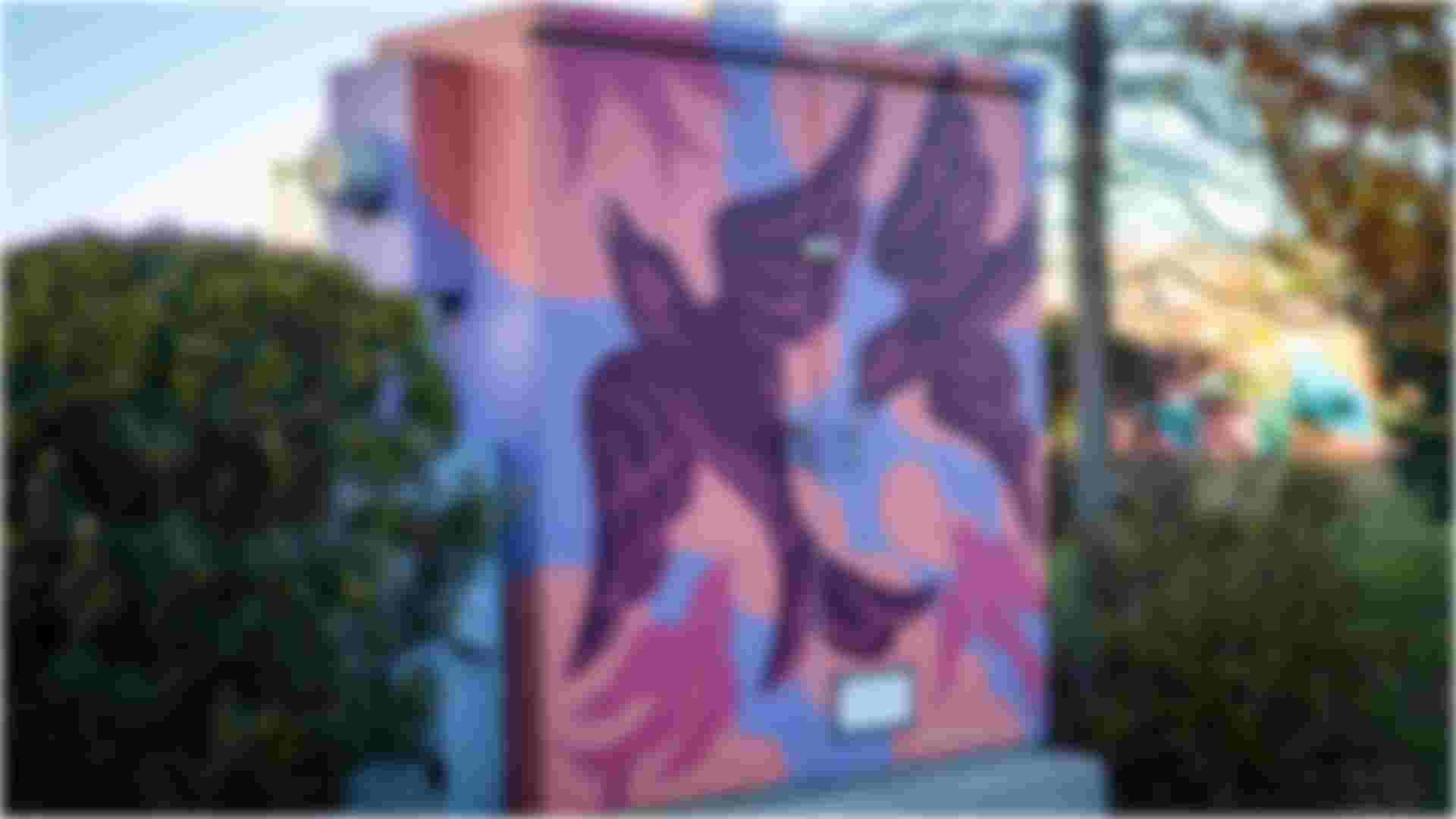 Painted utility box decorated with two stylized birds in flight, rendered in dark purple against a pastel background of pink, peach, and blue shapes, set among shrubs and trees.