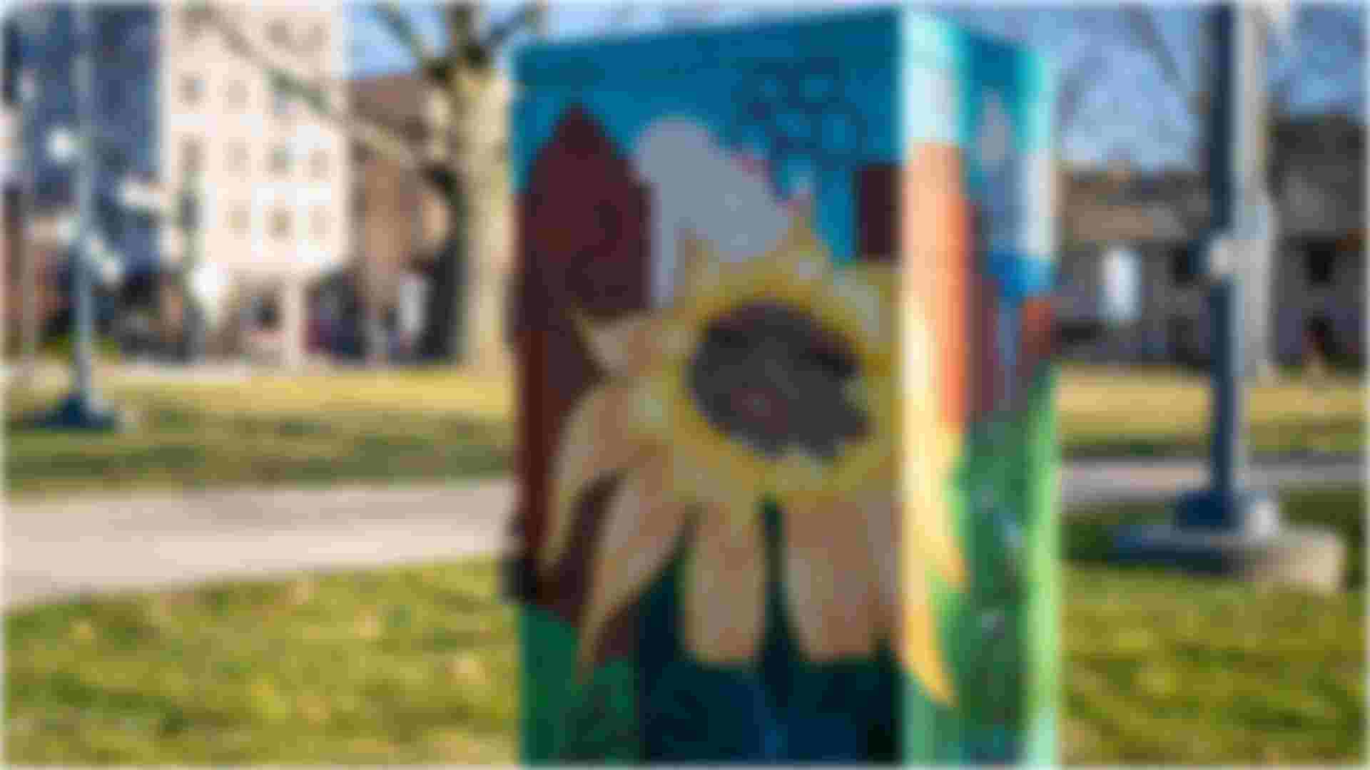 A painted utility box mural of a sunflower with a human face holding their knees in front of a city skyline and outlined flowers throughout.