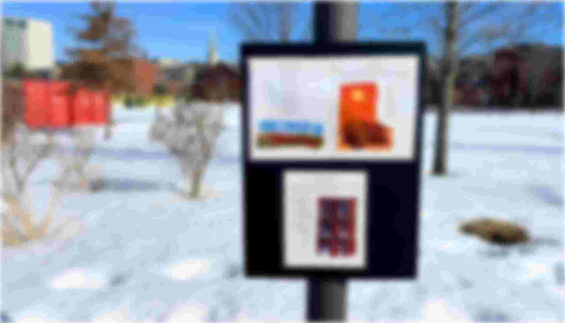 StoryWalk display sign in 195 District Park showing illustrated book pages mounted on a light post, surrounded by snow-covered ground, bare trees, and downtown Providence buildings under a clear blue winter sky.